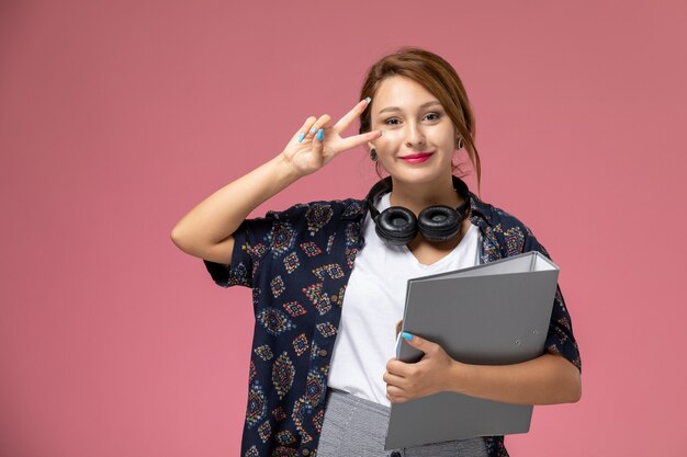 Giovane studentessa di vista frontale in maglietta bianca e pantaloni grigi che sorride con gli auricolari sull'istituto universitario di lezioni dello studente di sfondo rosa