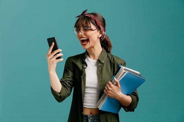 Giovane studentessa furiosa che indossa occhiali bandana con grandi blocchi di appunti con penna e telefono cellulare in un'altra mano guardando il telefono cellulare gridando ad alta voce isolato su sfondo blu