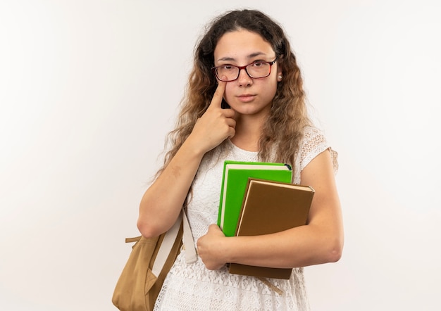 Giovane studentessa graziosa con gli occhiali e borsa posteriore che tengono i libri e mettendo il dito sotto gli occhi guardando la telecamera isolata su sfondo bianco con spazio di copia