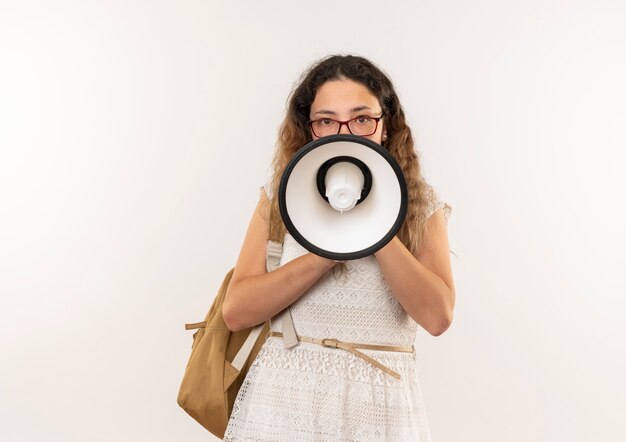 Giovane studentessa graziosa con gli occhiali e borsa posteriore parlando da altoparlante guardando la telecamera isolata su sfondo bianco con spazio di copia