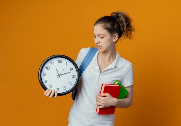 Giovane studentessa graziosa dispiaciuta che indossa la borsa posteriore che tiene e che guarda l'orologio e che tiene i libri isolati su fondo con lo spazio della copia