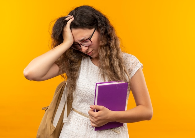 Giovane studentessa graziosa dolorante con gli occhiali e borsa posteriore che tiene il libro mettendo la mano sulla testa guardando verso il basso isolato su giallo con spazio di copia