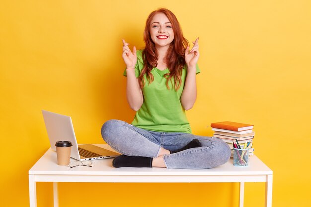 Giovane studentessa nel posto di lavoro con laptop e libri, seduta con le dita incrociate, augura il meglio, seduta con le gambe incrociate sul tavolo bianco, guarda sorridendo direttamente alla telecamera.