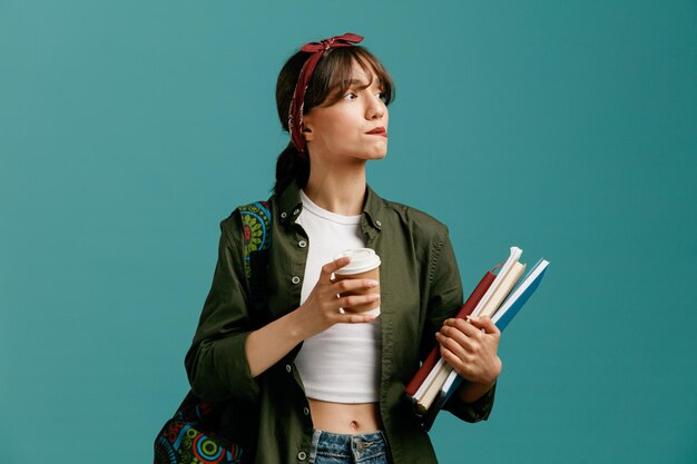 Giovane studentessa premurosa che indossa bandana e zaino con taccuini e tazza di caffè di carta guardando di lato mentre si morde il labbro isolato su sfondo blu