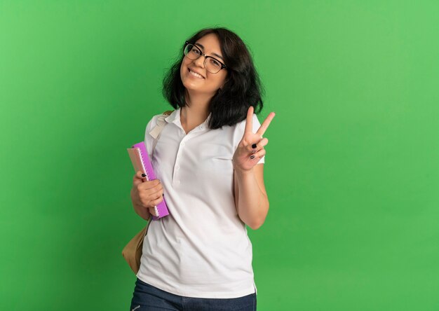 Giovane studentessa sorridente piuttosto caucasica con gli occhiali e borsa posteriore gesti il segno della mano di vittoria che tiene i libri sul verde con lo spazio della copia