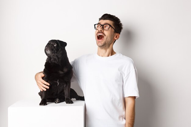 Giovane stupito con gli occhiali che abbraccia il suo cane, il proprietario dell'animale domestico e il carlino che fissano l'offerta promozionale nell'angolo in alto a sinistra, in piedi su sfondo bianco.