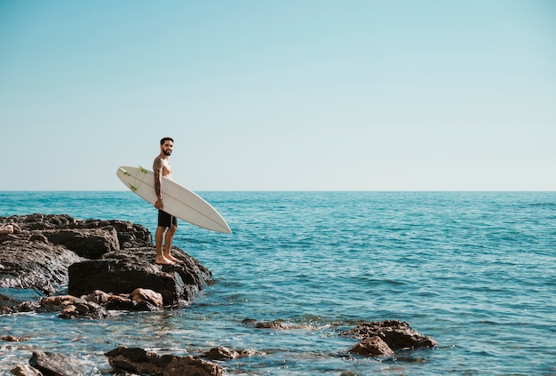 Giovane surfista in piedi sulla riva rocciosa