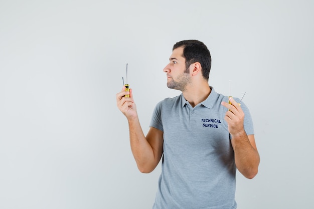 Giovane tecnico in uniforme grigia che tiene i cacciaviti in entrambe le mani e che osserva concentrato.