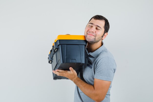 Giovane tecnico in uniforme grigia che tiene la cassetta degli attrezzi con entrambe le mani, sorridendo e guardando ottimista.