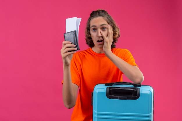 Giovane tipo bello in maglietta arancio che sta con i biglietti di aria della tenuta della valigia di viaggio che sembrano stanchi e annoiati