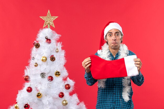 Giovane triste emotivo con il cappello di Babbo Natale in una camicia blu spogliata e che tiene il calzino di Natale