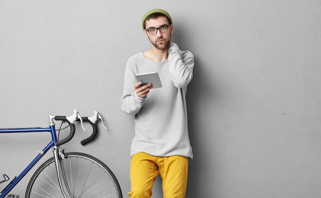 Giovane turista barbuto che tiene in mano una tavoletta moderna, alla ricerca di un percorso dove andare e cosa esplorare dopo con la sua bicicletta. Ragazzo alla moda con gadget moderni a casa prima di andare in bicicletta