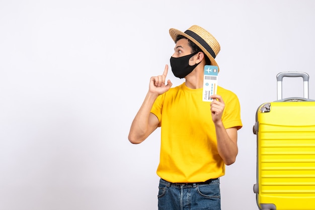 Giovane turista di vista frontale con il cappello di paglia che sta vicino alla valigia gialla che tiene il biglietto aereo
