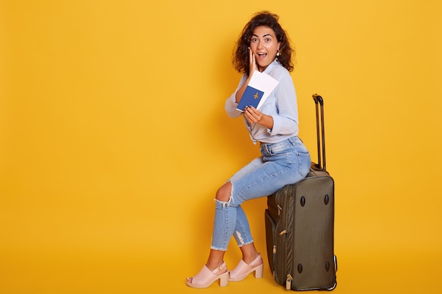 Giovane turista femminile allegro allegro emozionante e felice che si siede sulla sua grande valigia