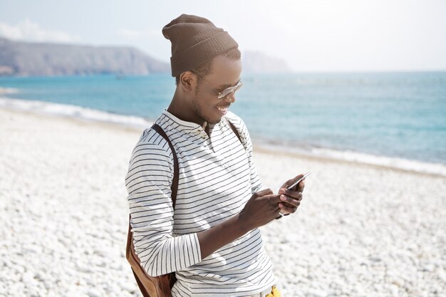 Giovane turista maschio europeo nero sorridente in cappello e tonalità usando Internet 3g sul cellulare sulla spiaggia, condividendo le immagini tramite i social media, godendo giorni felici durante le sue vacanze estive al mare