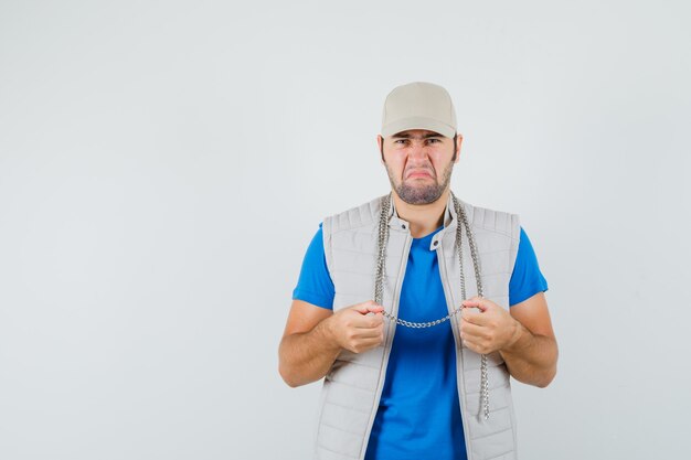 Giovane uomo accigliato mentre si tiene la collana a catena in t-shirt, giacca, berretto vista frontale.