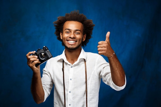 Giovane uomo africano bello che tiene la vecchia macchina fotografica su sfondo blu