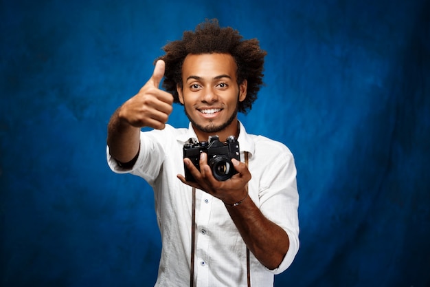 Giovane uomo africano bello che tiene vecchia macchina fotografica sopra la parete blu.