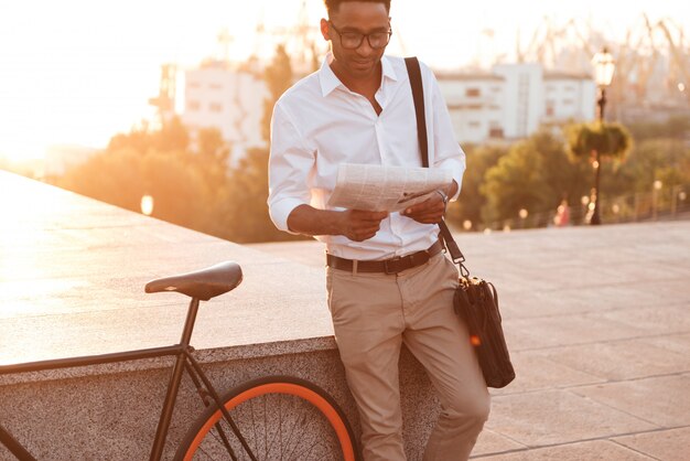 Giovane uomo africano bello nel primo mattino con il giornale della lettura della bicicletta.
