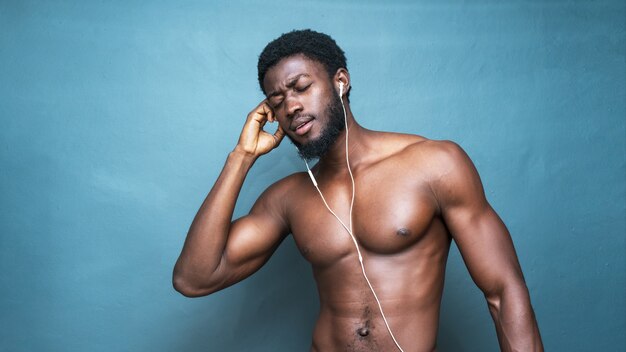 Giovane uomo africano in topless caldo che ascolta musica con gli auricolari su un blu