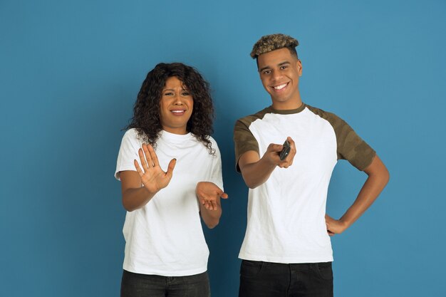 Giovane uomo afro-americano emotivo e donna in abiti casual bianchi in posa su sfondo blu. Bella coppia. Concetto di emozioni umane, espansione facciale, relazioni, annuncio. Guarda la TV insieme.