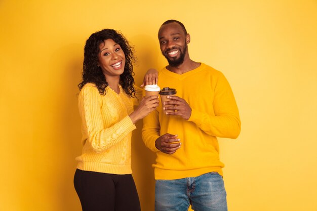 Giovane uomo afro-americano emotivo e donna in abiti casual luminosi su sfondo giallo. Bella coppia. Concetto di emozioni umane, espansione facciale, relazioni, annuncio. Bere il caffè insieme.