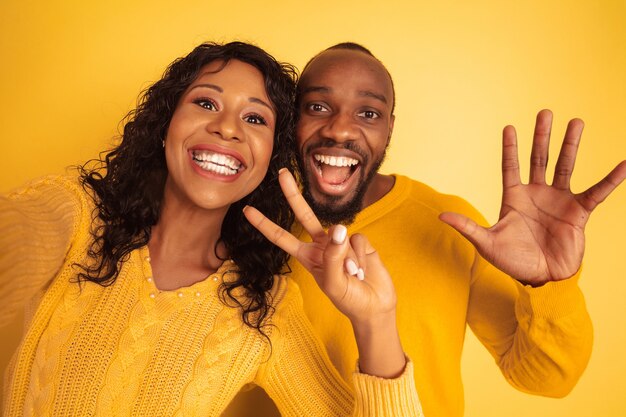 Giovane uomo afro-americano emotivo e donna in abiti casual luminosi su sfondo giallo. Bella coppia. Concetto di emozioni umane, espansione facciale, relazioni, annuncio. Facendo selfie insieme.
