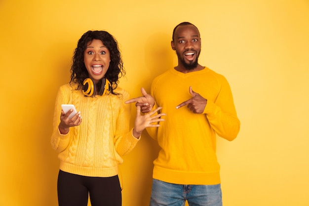 Giovane uomo afro-americano emotivo e donna in abiti casual luminosi su sfondo giallo. Bella coppia. Concetto di emozioni umane, espansione facciale, relazioni. Puntamento scioccato sullo smartphone.