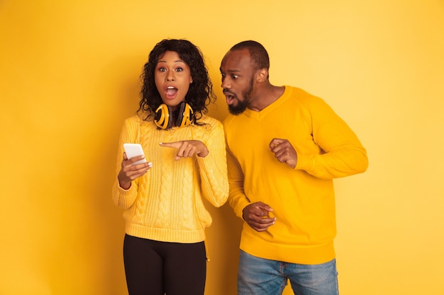 Giovane uomo afro-americano emotivo e donna in abiti casual luminosi su sfondo giallo. Bella coppia. Concetto di emozioni umane, espansione facciale, relazioni. Puntamento scioccato sullo smartphone.