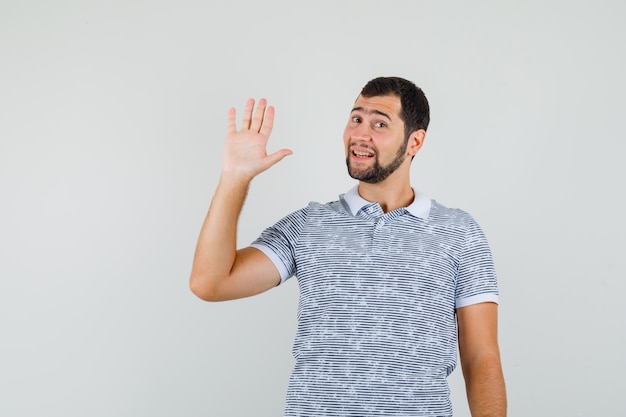 Giovane uomo agitando la mano per il saluto in t-shirt e guardando allegro, vista frontale.
