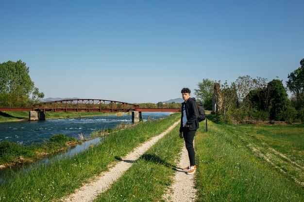 Giovane uomo alla moda che si leva in piedi sulla via vicino al fiume con lo zaino di trasporto