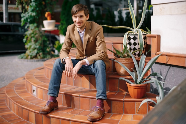 Giovane uomo alla moda sorridente che si siede all'aperto sulle scale circolari d'annata