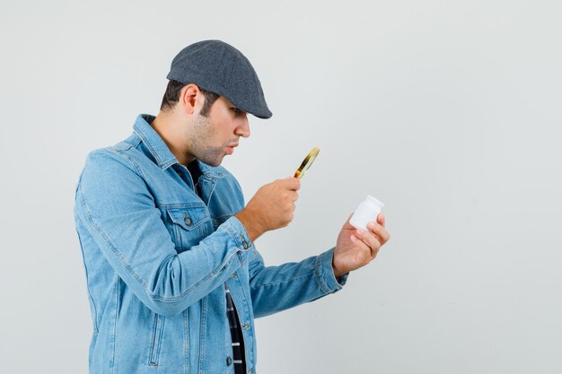 Giovane uomo alla ricerca di una bottiglia di pillole con lente d'ingrandimento in giacca, berretto e guardando concentrato.