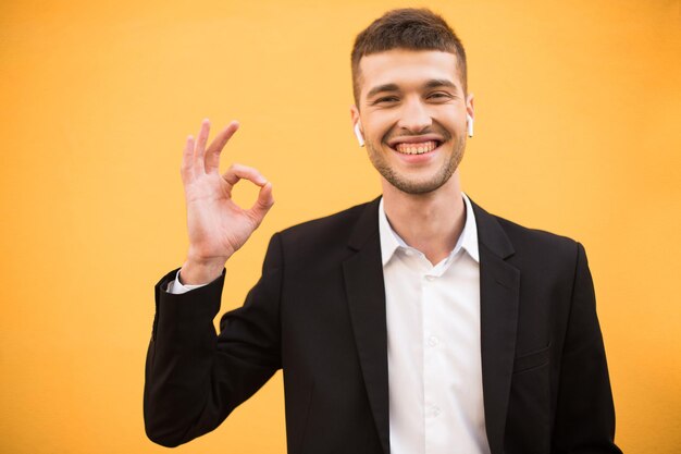 Giovane uomo allegro nella classica giacca nera e camicia bianca che mostra felicemente il gesto giusto mentre guarda nella fotocamera su sfondo arancione