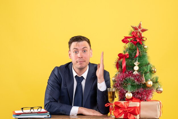 giovane uomo alzando la mano seduto al tavolo vicino albero di Natale e regali su giallo
