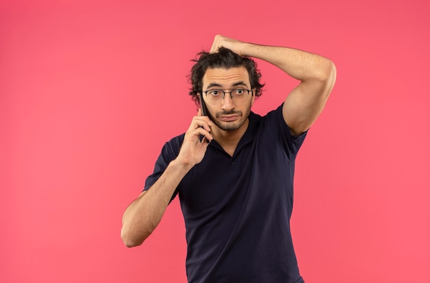 Giovane uomo ansioso in camicia nera con vetri ottici parla al telefono e tiene la testa isolata sul muro rosa