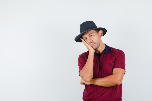 Giovane uomo appoggiato guancia sul palmo alzato in t-shirt, cappello e guardando assonnato, vista frontale.