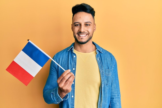 Giovane uomo arabo che tiene la bandiera della francia che sembra positiva e felice in piedi e sorridente con un sorriso fiducioso che mostra i denti