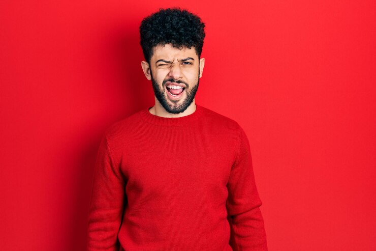 Foto gratuito giovane uomo arabo con la barba che indossa un maglione rosso casual che strizza l'occhio guardando la telecamera con espressione sexy, faccia allegra e felice.
