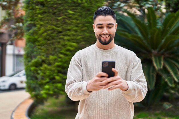 Giovane uomo arabo sorridente fiducioso utilizzando lo smartphone al parco