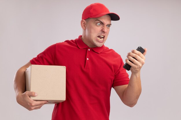 Giovane uomo arrabbiato di consegna che indossa l'uniforme con la scatola della tenuta del cappuccio e che esamina il telefono nella sua mano isolata sulla parete bianca