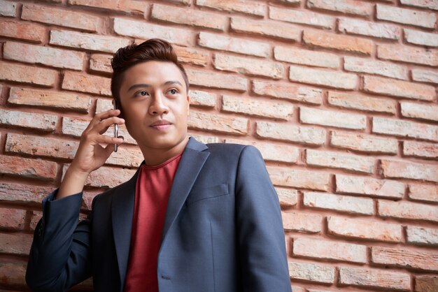 Giovane uomo asiatico alla moda che sta davanti al muro di mattoni e che tiene smartphone all'orecchio