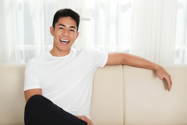 Giovane uomo asiatico allegro che si siede sullo strato a casa
