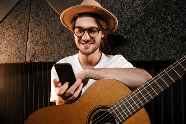 Giovane uomo attraente con la chitarra che utilizza felicemente lo smartphone nel moderno studio di registrazione del suono