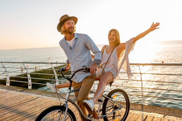 Giovane uomo attraente e donna che viaggiano in bicicletta, coppia romantica in vacanza estiva in riva al mare al tramonto, vestito stile boho hipster, amici che si divertono insieme