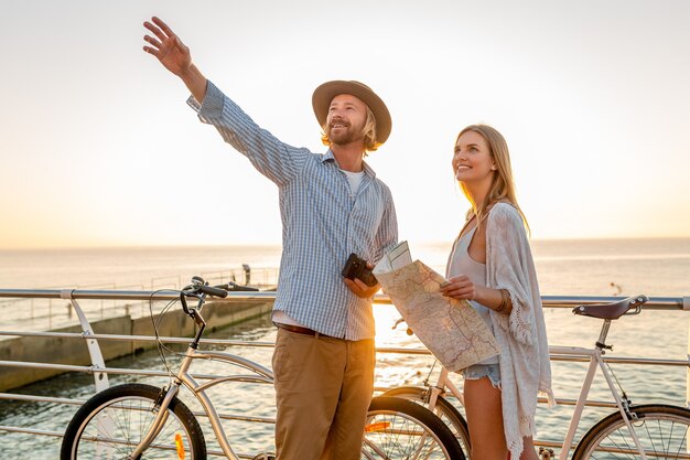 Giovane uomo attraente e donna che viaggiano in bicicletta, tenendo mappa e visite turistiche, coppia romantica in vacanza estiva in riva al mare al tramonto, amici che si divertono insieme