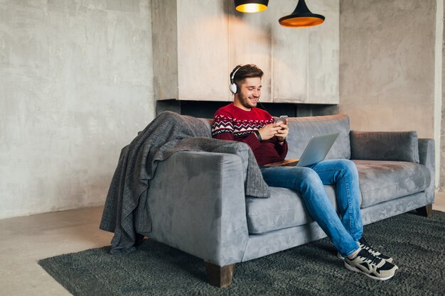 Giovane uomo attraente sul divano a casa in inverno con lo smartphone in cuffia, ascoltando musica, indossando un maglione lavorato a maglia rosso, lavorando sul computer portatile, libero professionista, sorridente, felice, positivo