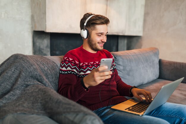 Giovane uomo attraente sul divano a casa in inverno con lo smartphone in cuffia, ascolto di musica, indossa un maglione lavorato a maglia rosso, lavora al computer portatile, libero professionista, sorridente, felice, positivo, digitando