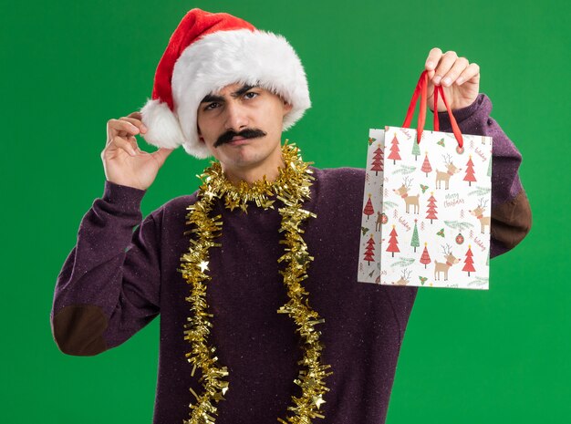 Giovane uomo baffuto che indossa il cappello di Babbo Natale con orpelli intorno al collo tenendo il sacchetto di carta con un regalo di Natale guardando la telecamera confuso e dissolto in piedi su sfondo verde