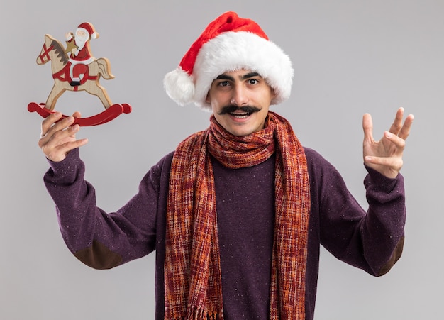Giovane uomo baffuto che indossa il cappello di Babbo Natale con sciarpa calda intorno al collo tenendo il giocattolo di Natale guardando la telecamera felice e allegro con il braccio alzato in piedi su sfondo bianco